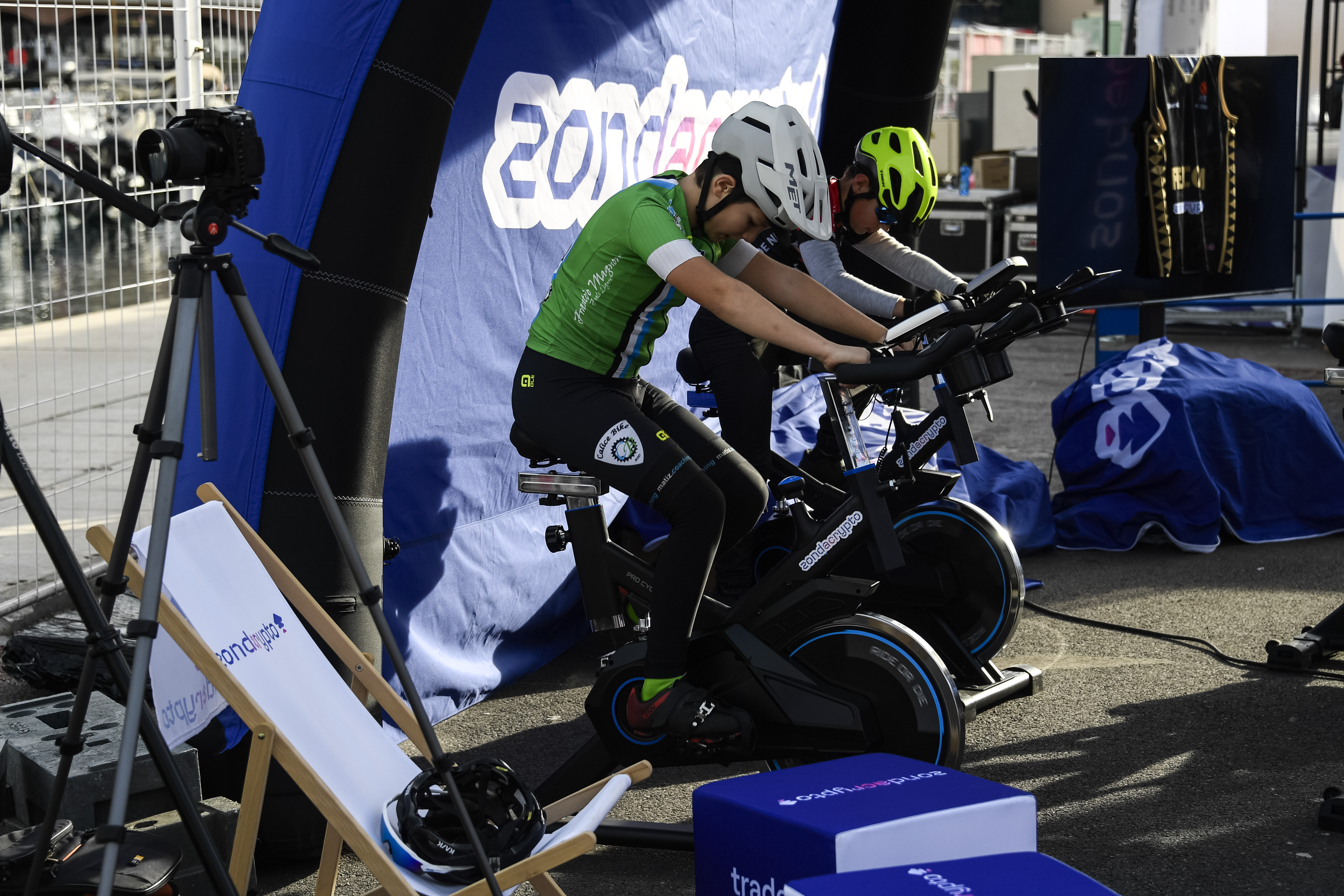 Two young cyclists on indoor bike trainers, wearing athletic gear and helmets, in an outdoor warm-up or staging area in front of a blue banner.