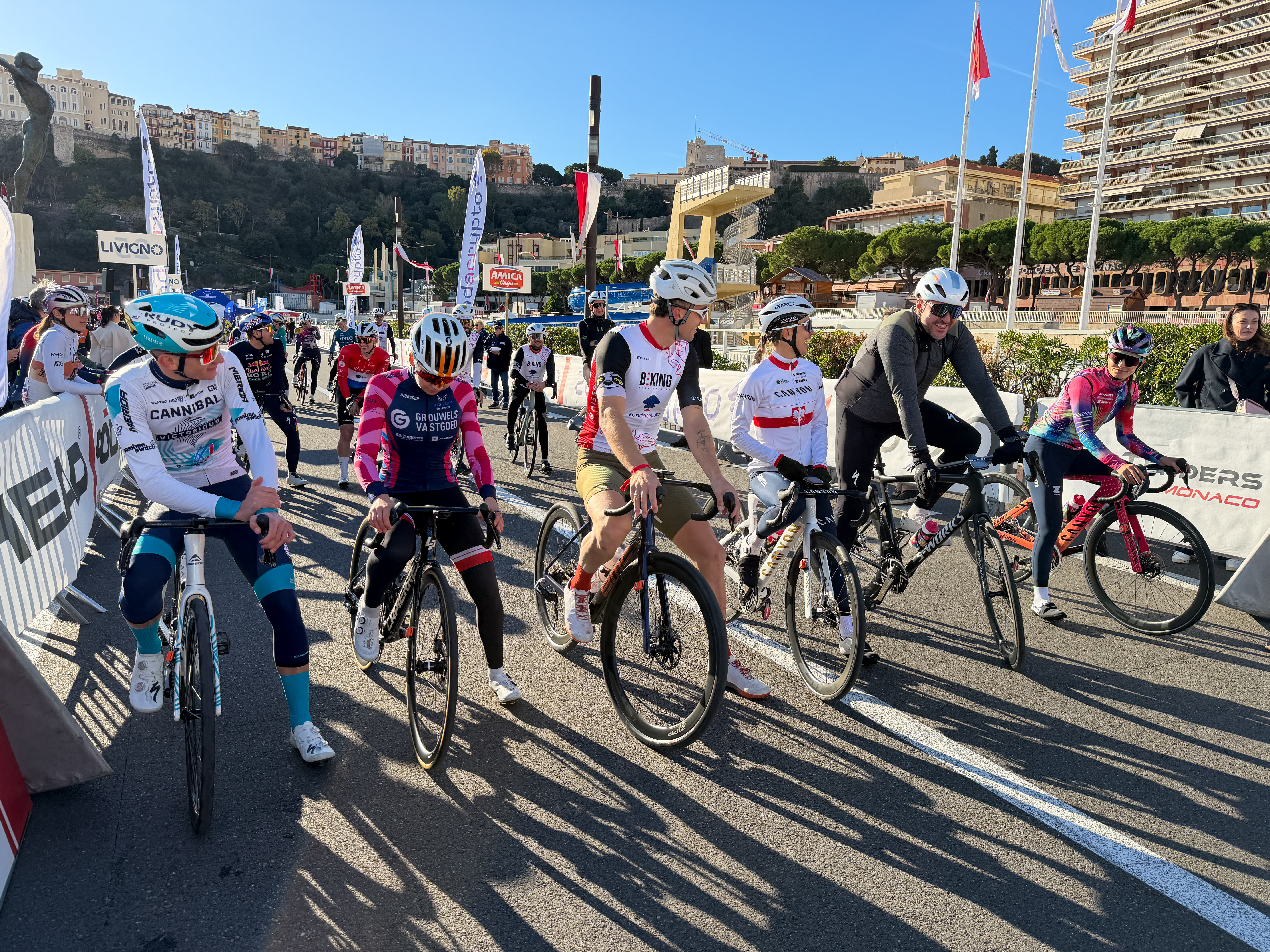A group of cyclists in colorful jerseys and helmets stands at the start line on a sunny street. Flags, stands, and city buildings on a hill are visible in the background.