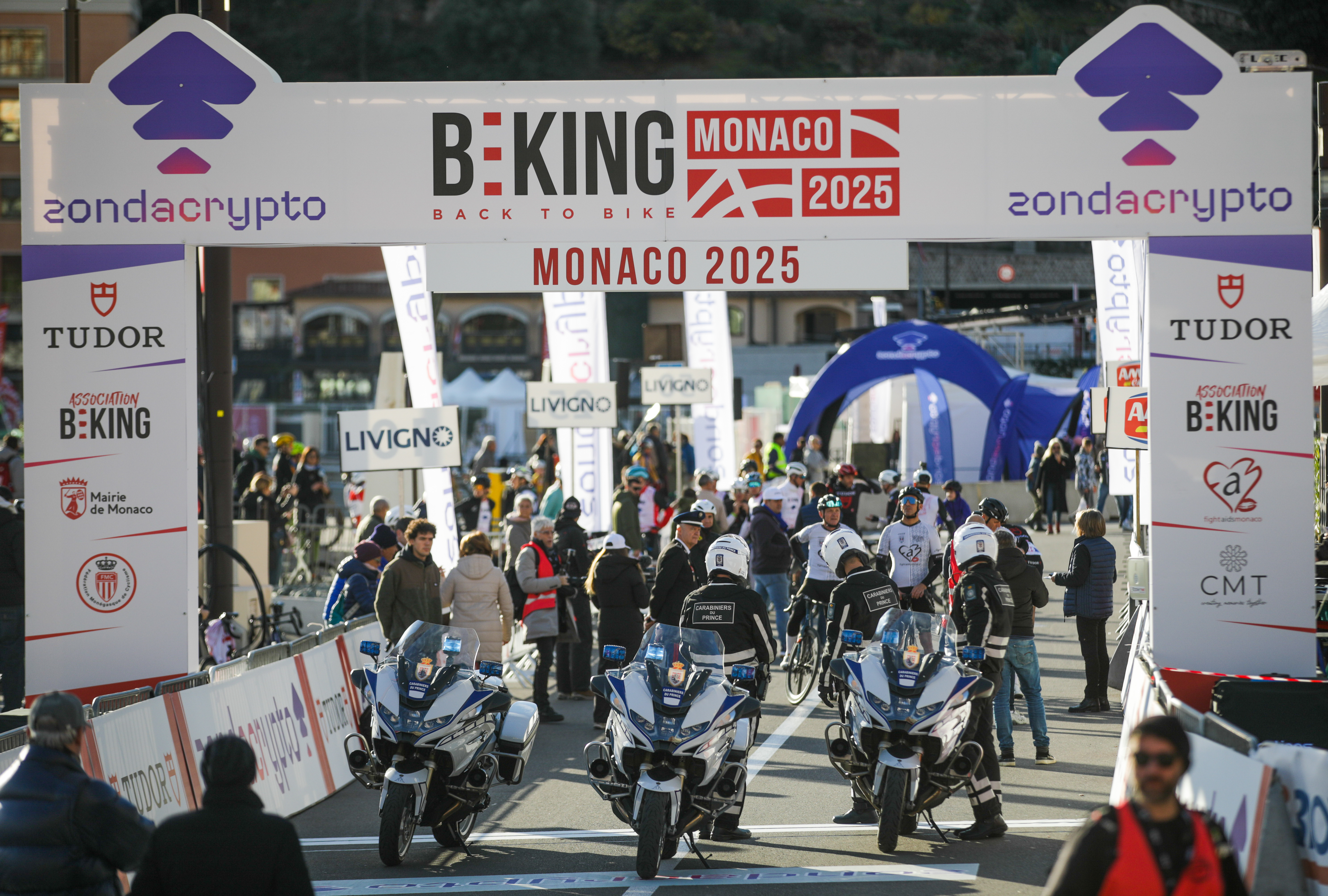 Three police motorcycles are parked beneath an archway displaying "B:KING MONACO 2025" and the "zondacrypto" logo. A crowd of people and cyclists is visible in the background along with sponsor advertisements.