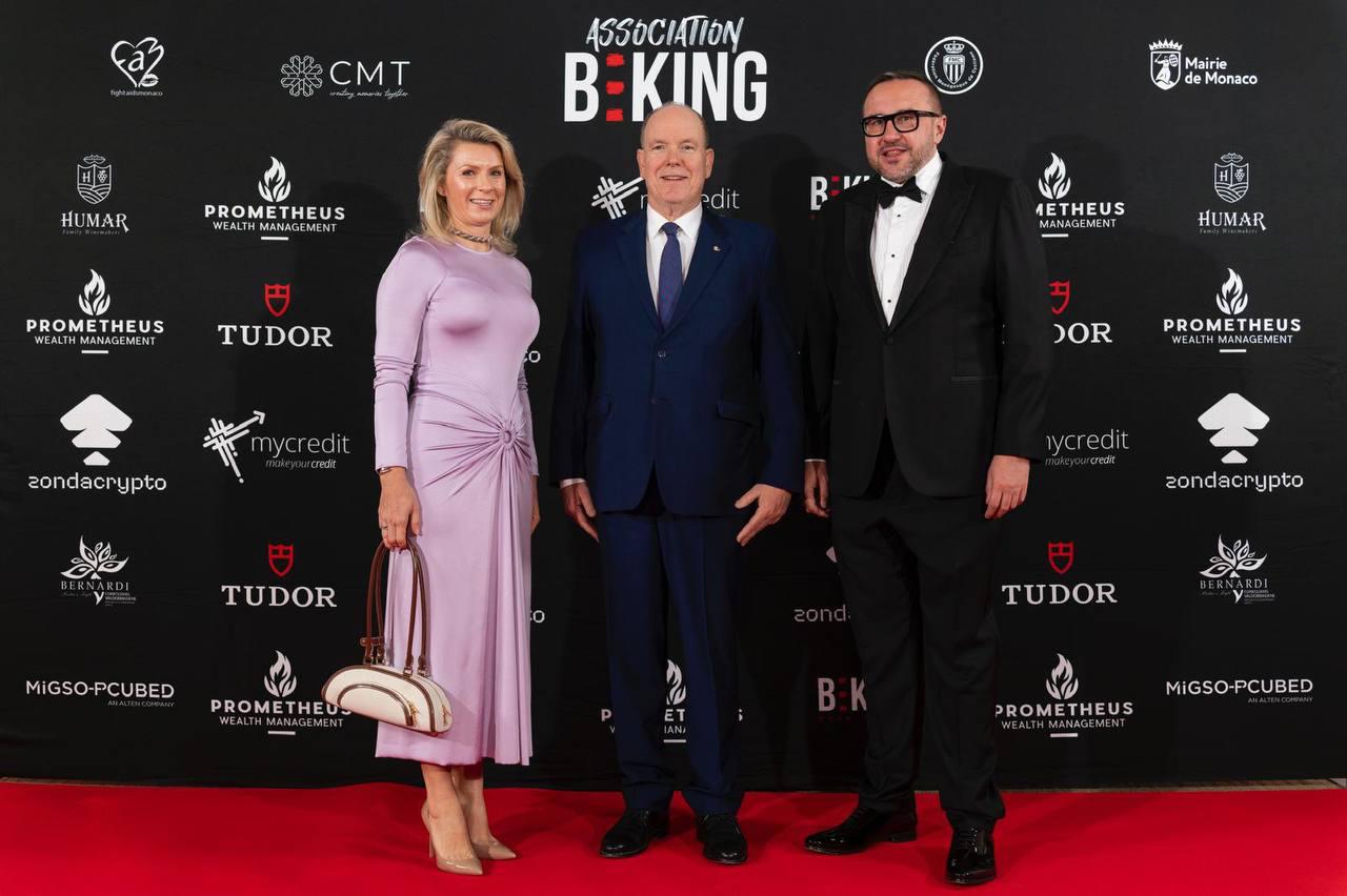 Three people — a woman in a fitted lilac dress, a man in a navy suit (Prince Albert II of Monaco), and a man in a black tuxedo with a bow tie — stand on a red carpet. The background is a sponsor wall with repeating company logos and the text "ASSOCIATION B:KING".