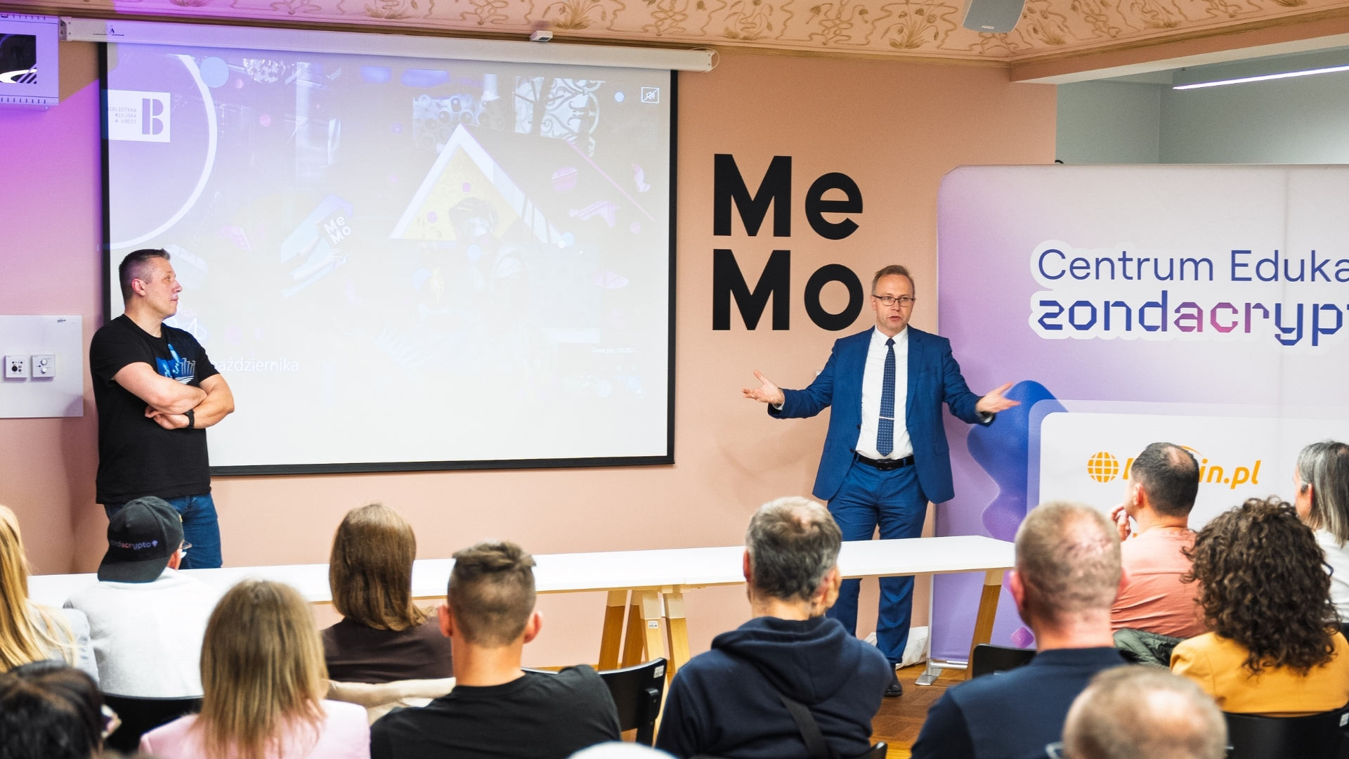 Mężczyzna w garniturze przemawia do publiczności podczas wydarzenia edukacyjnego o krypto, z inną osobą stojącą po lewej stronie i dużym bannerem zondacrypto 'Centrum Edukacji' w tle.