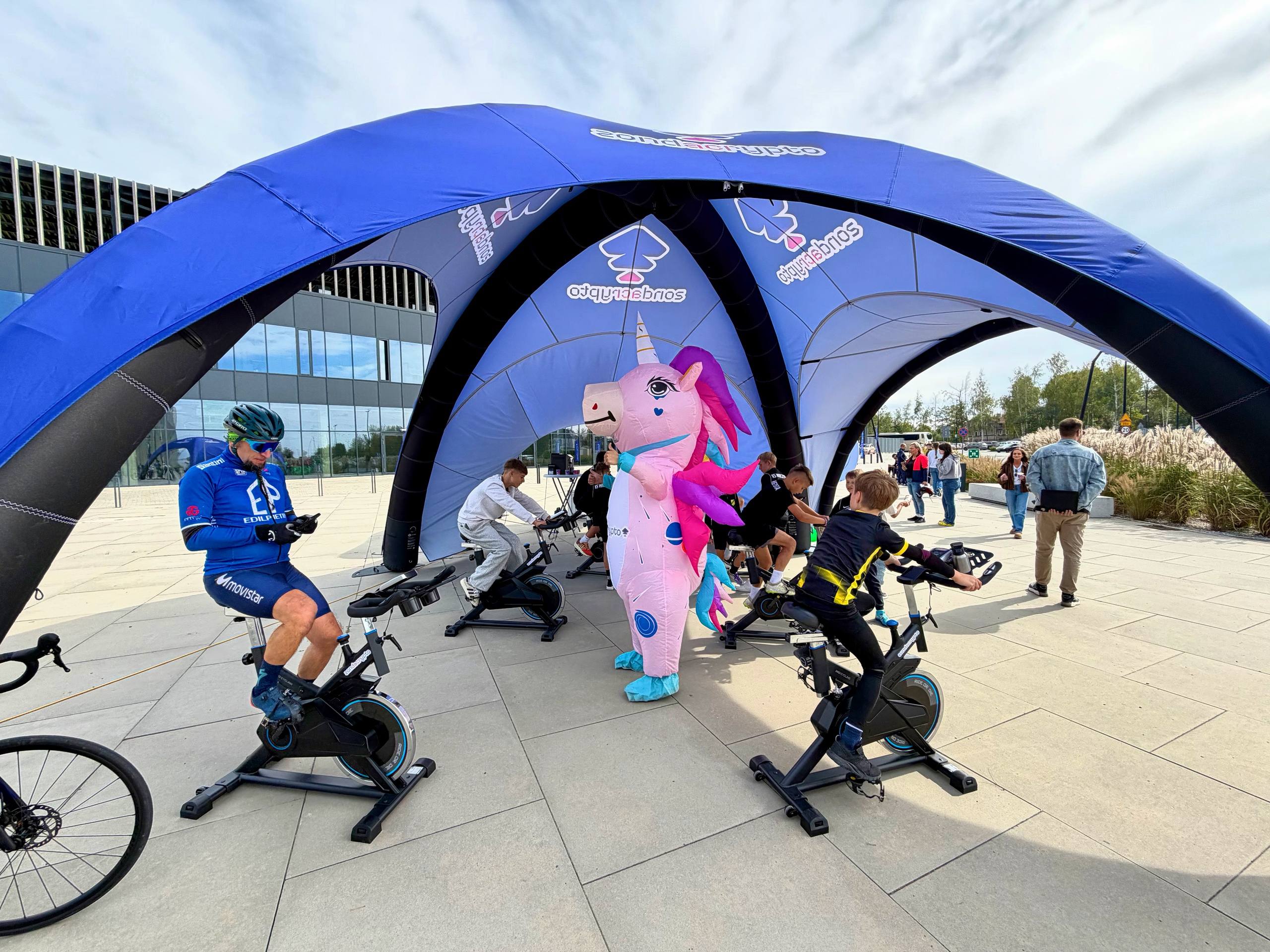 Outdoor scene at ArcelorMittal Park: blue zondacrypto inflatable tent with stationary bikes, children and an adult riding, Zondolo pink unicorn cheering; modern building in the background.