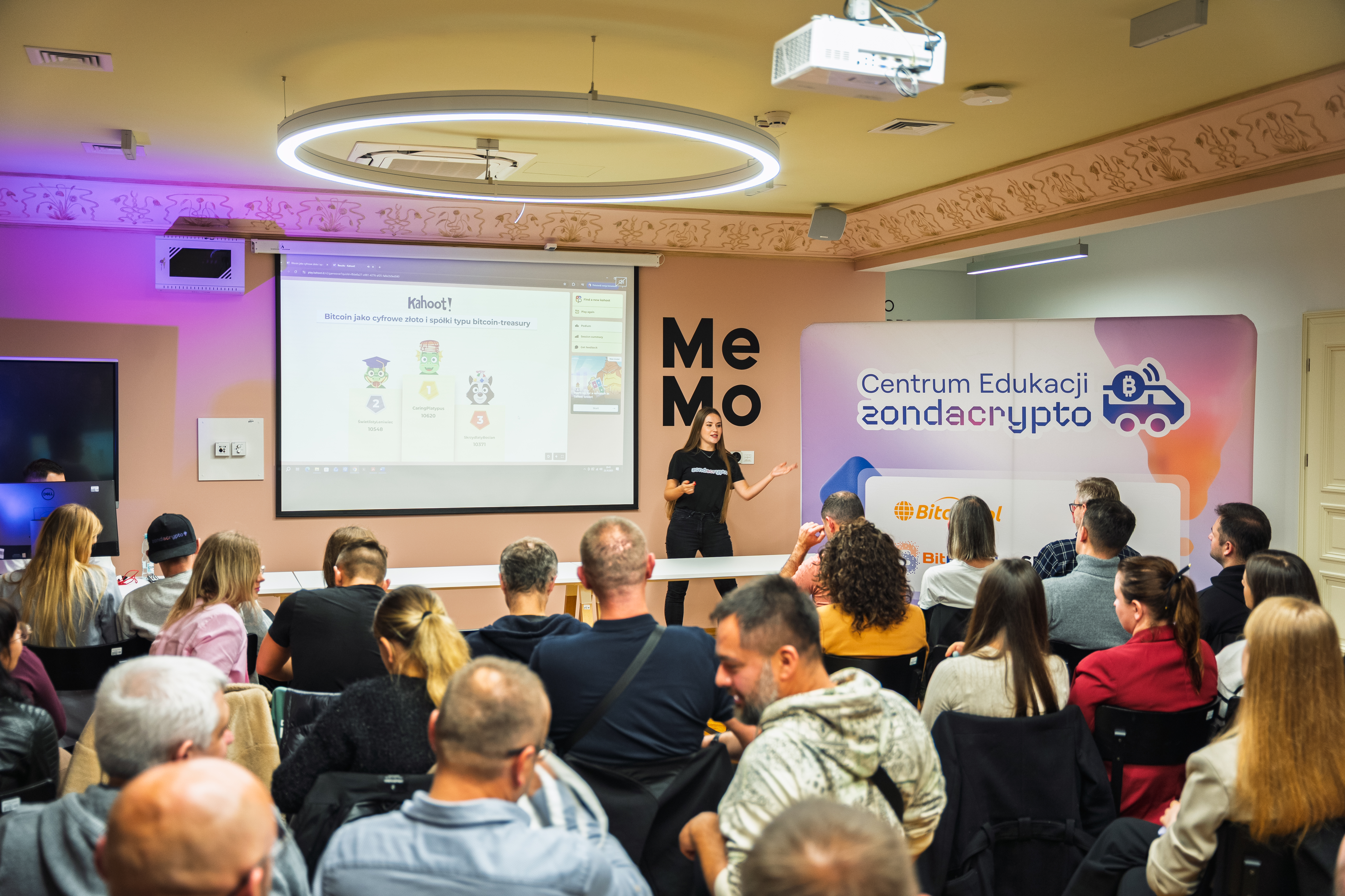 Prelegentka na scenie, stojąca przed ekranem projektora wyświetlającym slajd quizu Kahoot!, angażująca publiczność na warsztatach edukacyjnych zondacrypto dotyczących krypto i blockchain.